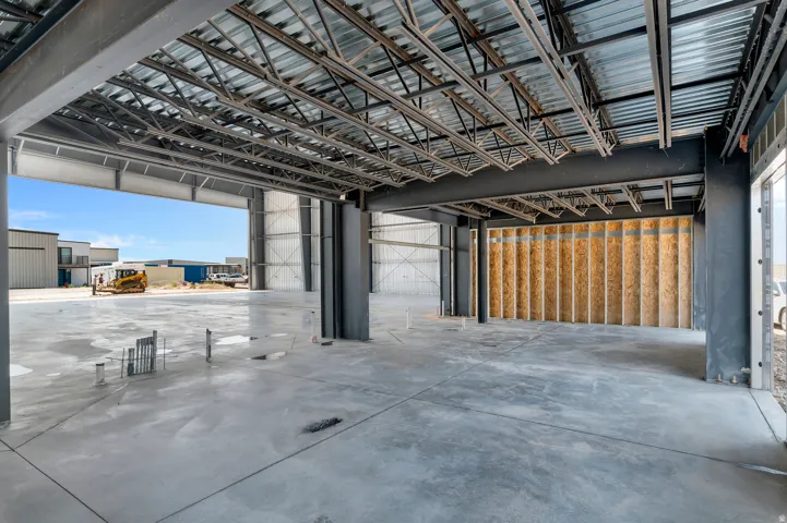 Spacious concrete slab flooring, exposed metal ceiling trusses, structural steel beams, unfinished wood-framed wall, and large overhead door opening
