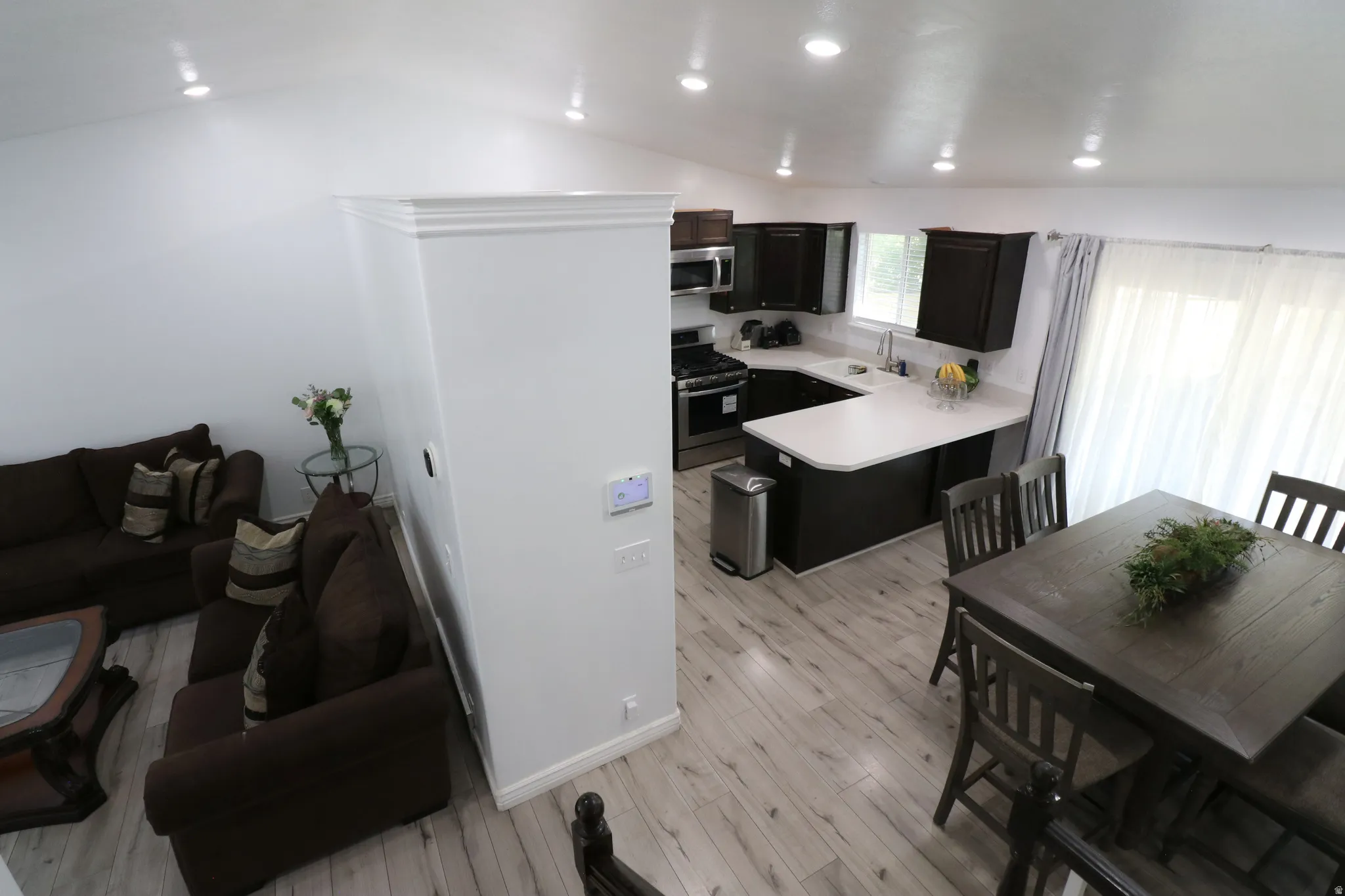 Open-concept living and dining area with wood-finish flooring throughout, modern black cabinetry and stainless steel appliances in the kitchen, white quartz countertops, recessed ceiling lighting, and large windows providing natural light