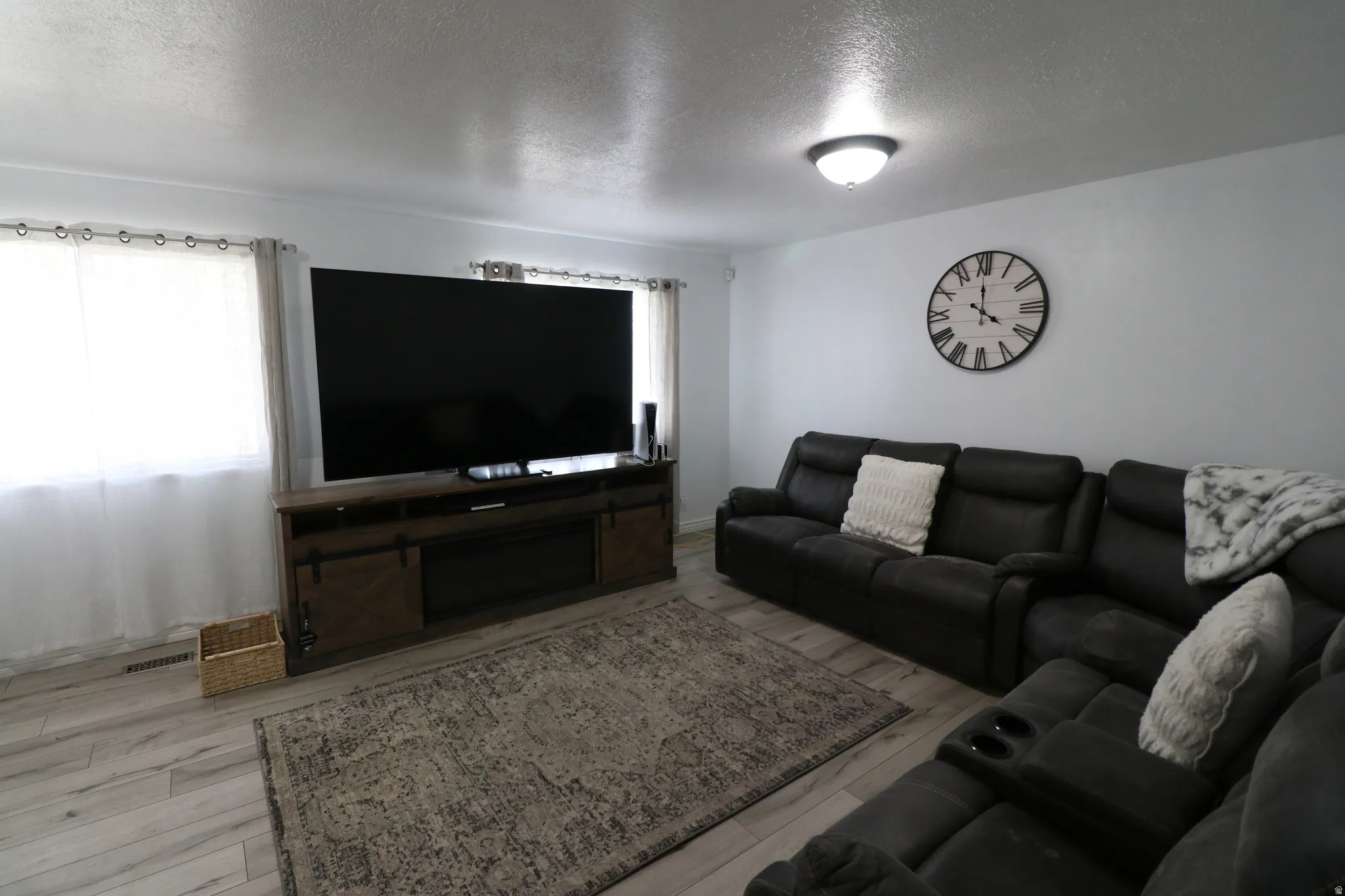 Spacious entertainment room featuring a large flat-screen television mounted above wood-finish media cabinetry, comfortable black leather sectional seating, wood-finish flooring throughout, neutral wall finishes, and recessed ceiling lighting with a wall-
