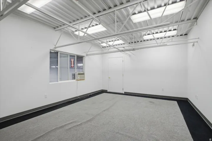 White painted walls, exposed metal ceiling joists, fluorescent overhead lighting, window with a wall-mounted air conditioning unit, and grey carpet flooring