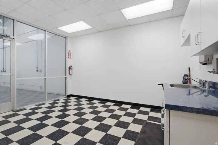 Modern commercial or institutional space featuring a striking black-and-white checkered tile floor, sleek glass partition walls with aluminum framing, bright overhead panel lighting, and a compact kitchenette with stainless steel sink and under-counter st