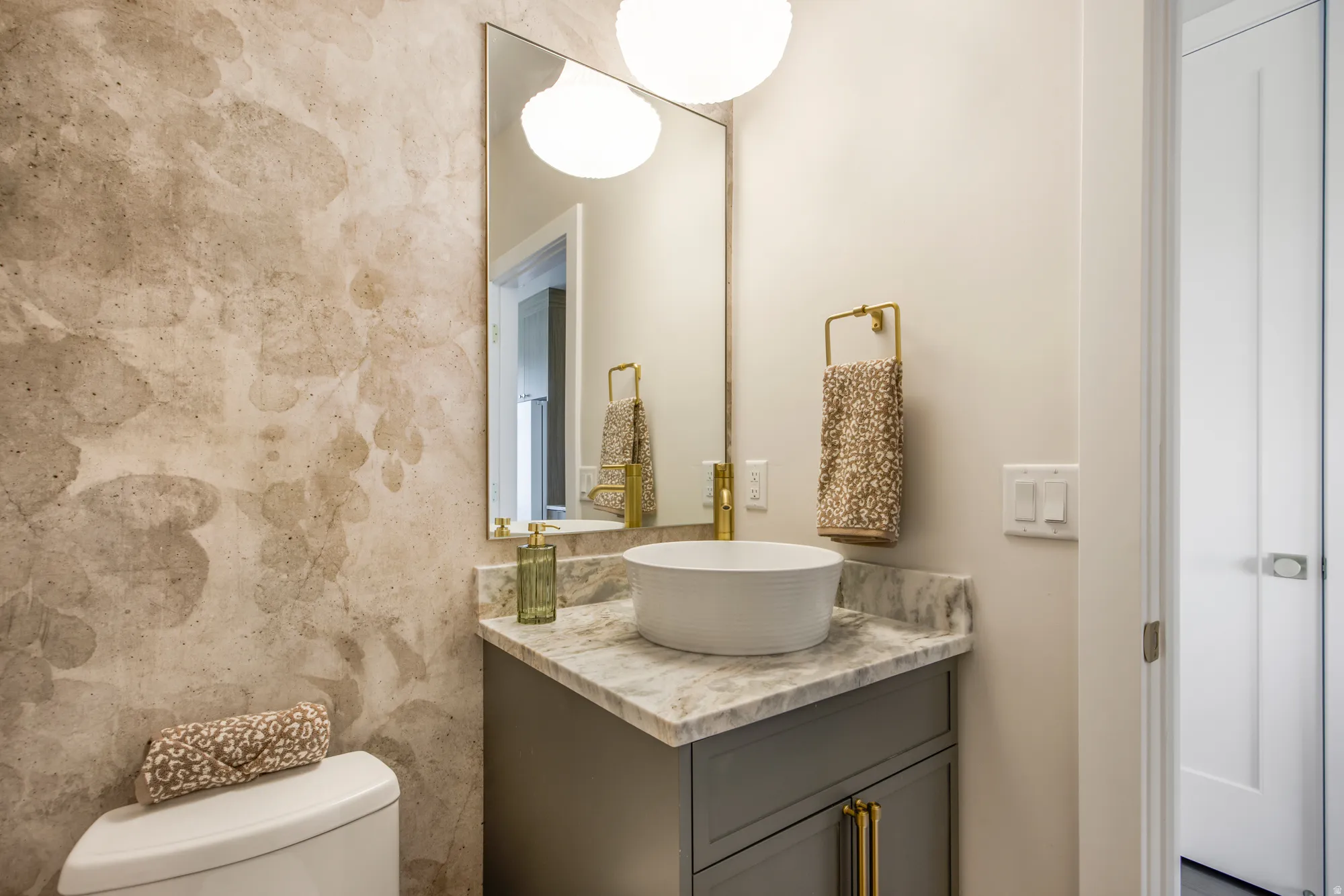 Textured wall covering, framed mirror, vanity with stone countertop, vessel sink, and gold-finish fixtures