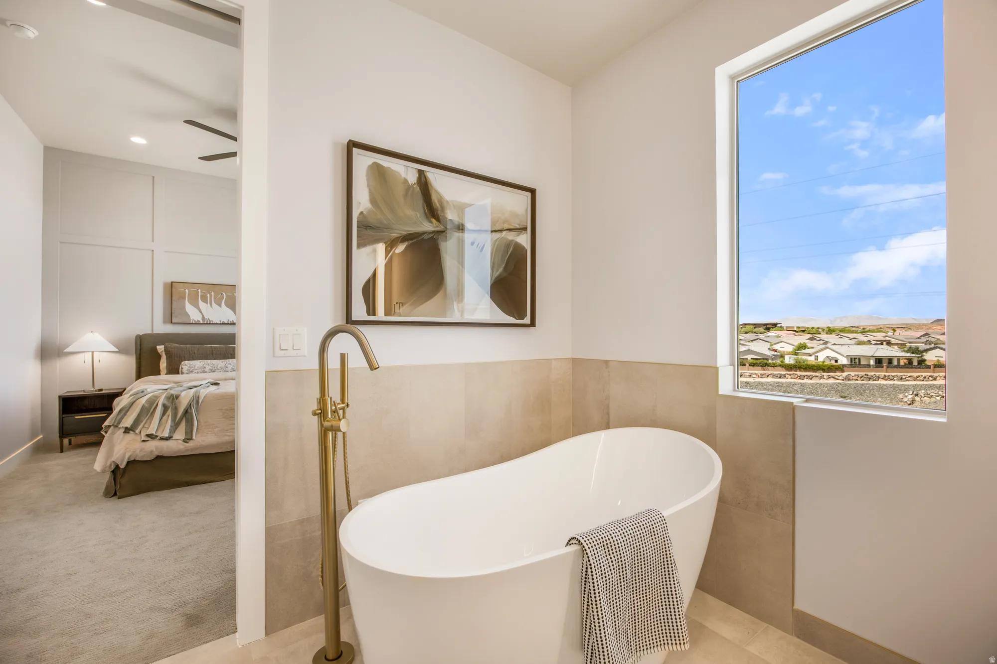 Freestanding soaking tub with a floor-mounted brass faucet, large window, and tile wainscoting