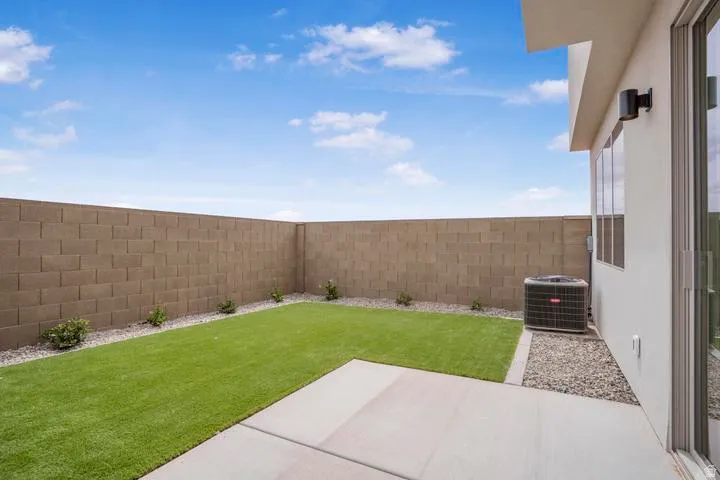 Spacious backyard oasis featuring a lush, manicured lawn bordered by composite block perimeter walls, a concrete patio extension, decorative gravel landscaping with accent plantings, and an air conditioning unit