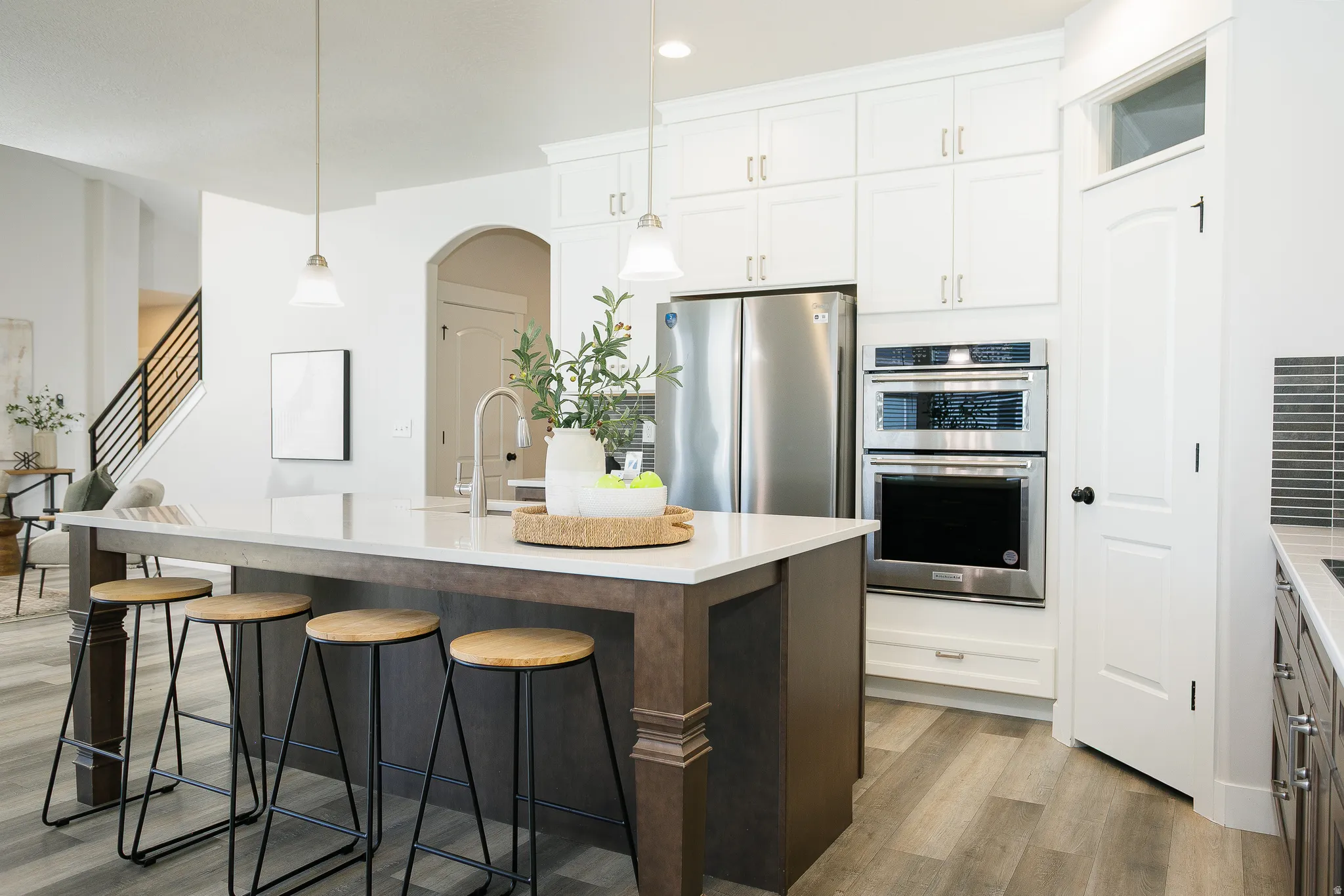Expansive kitchen island with seating, stainless steel appliances, built-in double ovens, white shaker cabinetry, and wood-finish flooring
