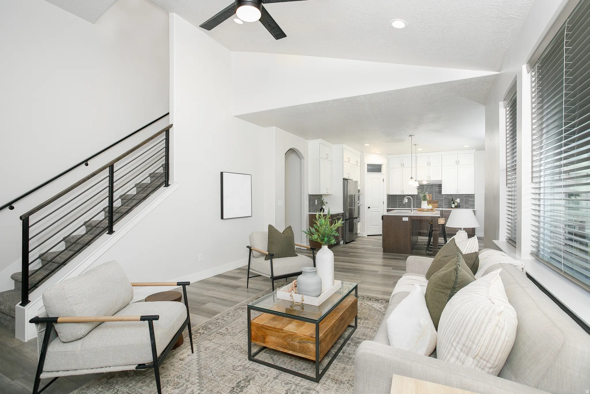 Open-concept living area featuring a modern black metal staircase railing, wood-finish flooring, white kitchen cabinetry with a dark backsplash, and large windows with blinds