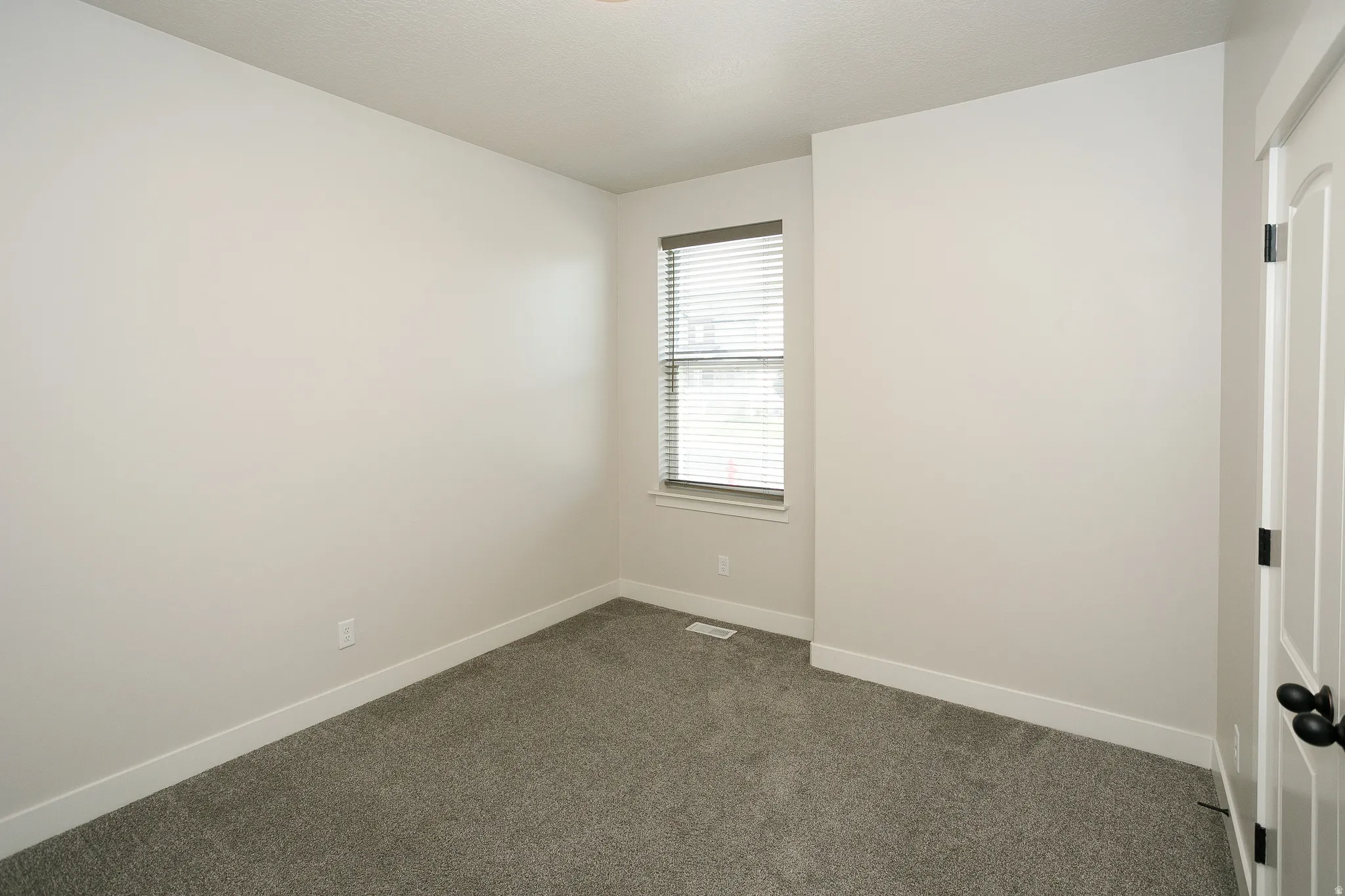 Neutral wall tones, window with horizontal blinds, textured carpet flooring, and a white interior door with black hardware