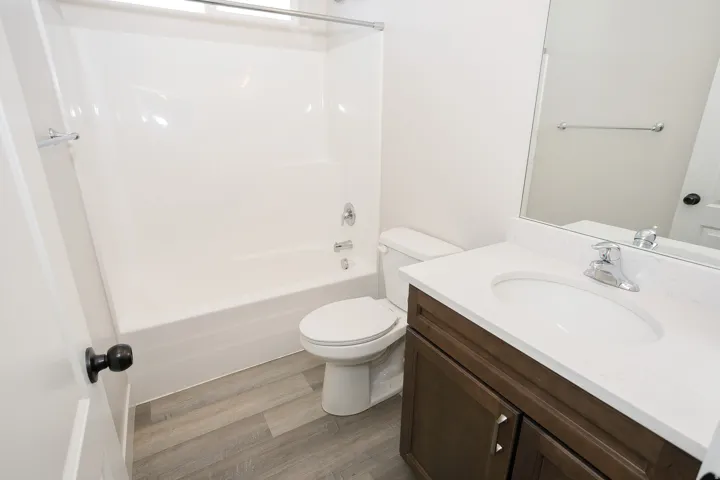 White bathtub with shower, white toilet, wood-finish flooring, vanity with white countertop, and dark wood-finish cabinetry