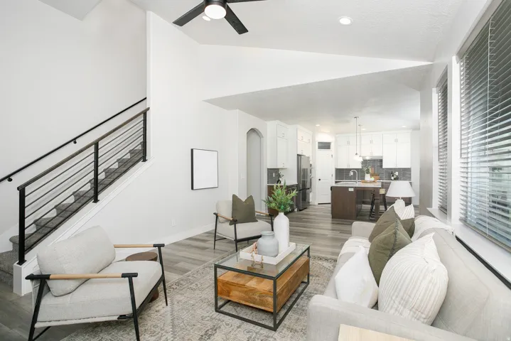 Open-concept living area featuring a modern black metal staircase railing, wood-finish flooring, white kitchen cabinetry with a dark backsplash, and large windows with blinds