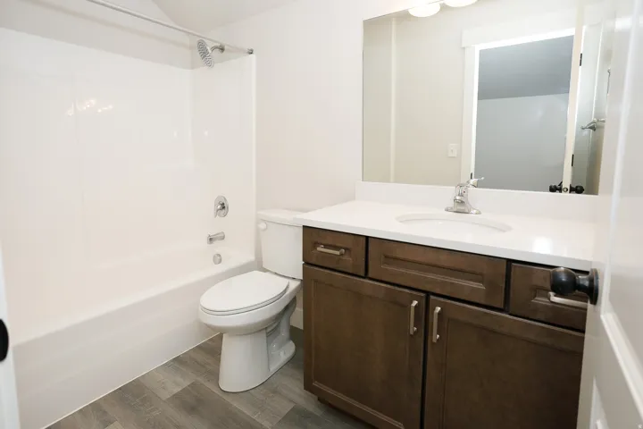 White bathtub and shower combination, wood-finish flooring, dark wood-finish vanity with drawer and cabinet storage, white countertop, and an integrated mirror
