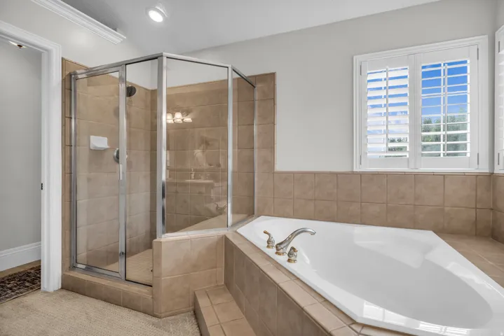 Primary bathroom featuring a soaking tub with deck-mounted faucet, frameless glass-enclosed shower with tile surround, neutral tile flooring, and natural light from dual casement windows with plantation shutters