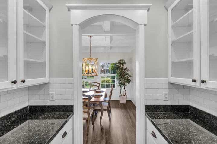 Elegant galley kitchen featuring white cabinetry with glass-front upper cabinets, dark stone countertops, subway tile backsplash, and an arched passageway framing the adjoining dining area
