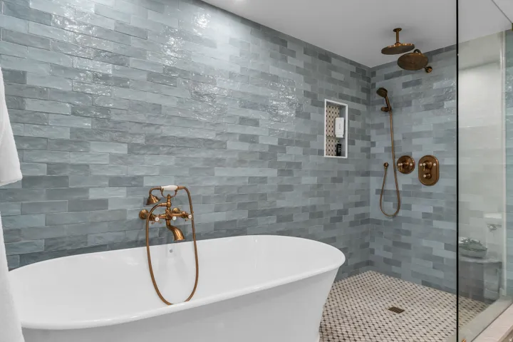 Basement Spa-inspired primary bathroom featuring a freestanding soaking tub, linear  tile wall treatment with varied finishes, brass-fixture rainfall showerhead and handheld spray, frameless glass shower enclosure, and geometric mosaic floor tile