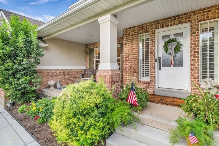Welcoming front entry featuring a brick exterior, white-trimmed covered porch with a support column, and manicured landscaping including vibrant shrubs and seasonal plantings