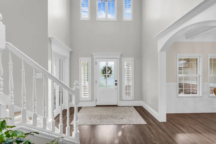 Welcoming foyer showcasing soaring ceilings with transom windows, elegant white balustraded staircase with turned spindles, wide-plank wood-finish flooring, and paneled front entry door flanked by sidelights with operable shutters