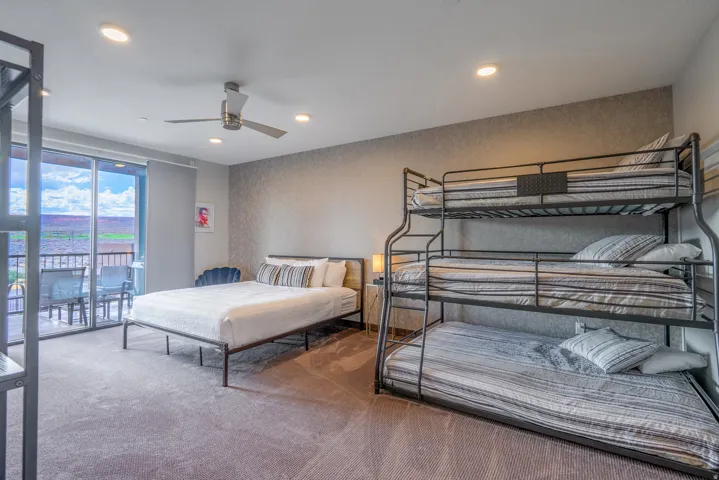 Spacious multi-bed dormitory-style room featuring three metal bunk bed units with integrated storage shelving, wood-finish flooring, recessed and ceiling fan lighting, and direct access to a balcony with water views