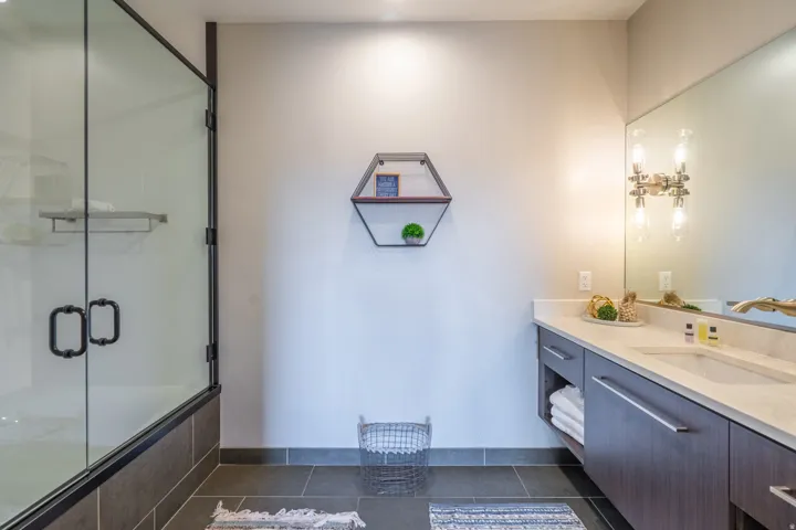 Walk-in shower with black-framed glass enclosure, dark gray tile flooring, wood-finish vanity with an undermount sink, light-toned countertop, and a wide wall mirror