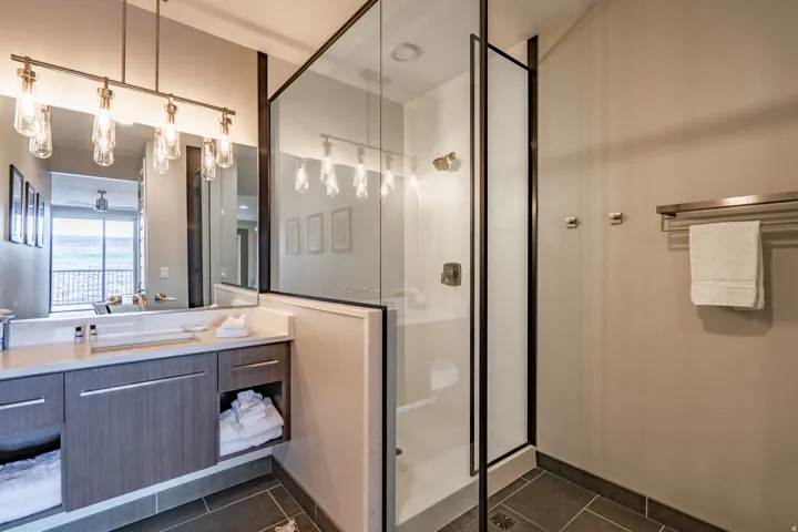 Floating vanity with integrated rectangular sink and quartz countertop, complemented by a glass-enclosed shower with dark-framed door, a multi-bulb vanity light fixture, and dark floor tile