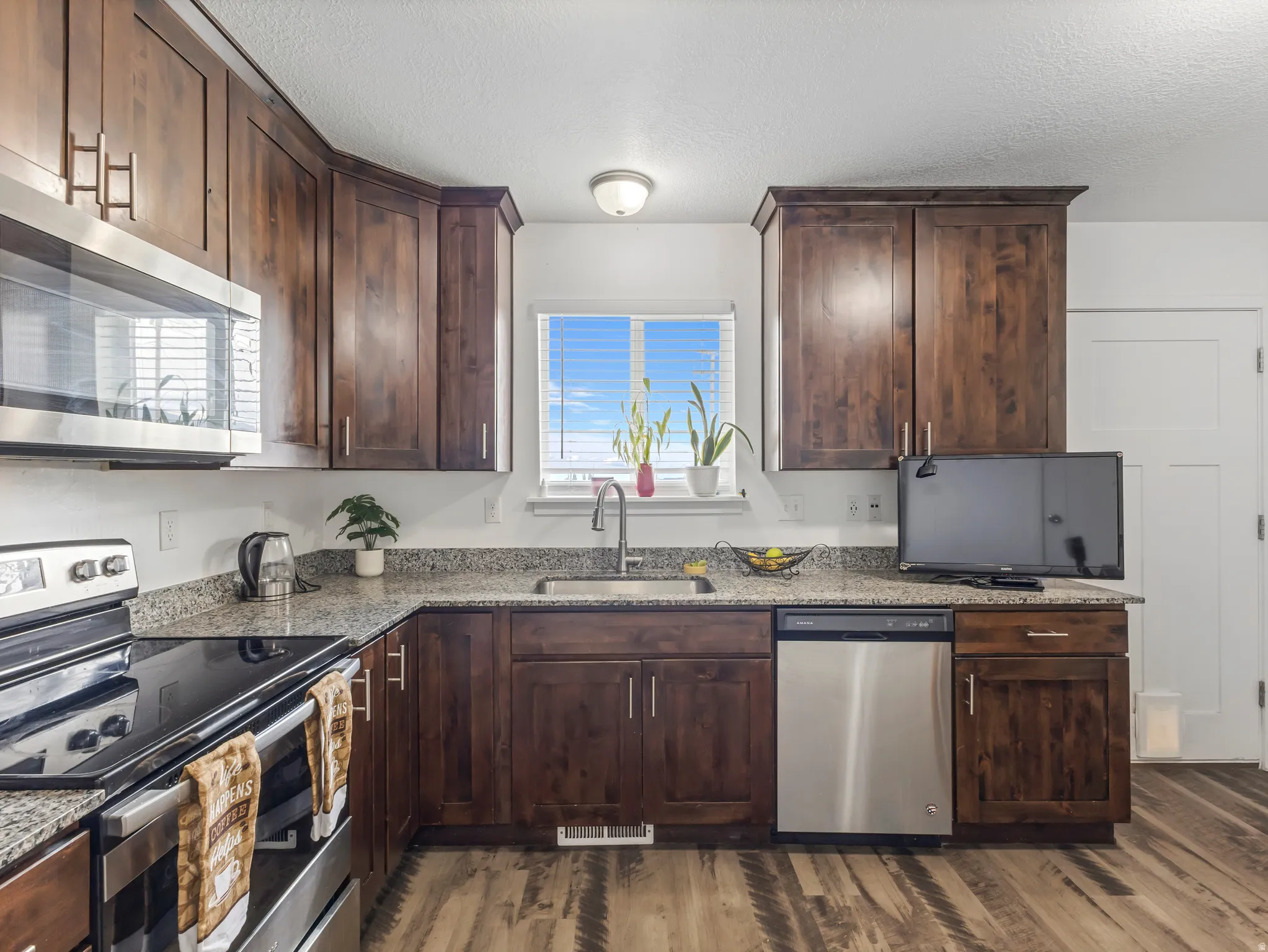 Kitchen featuring wood-finish flooring, granite-style countertops, stainless steel appliances, dark wood cabinetry, and a stainless steel sink with a gooseneck faucet