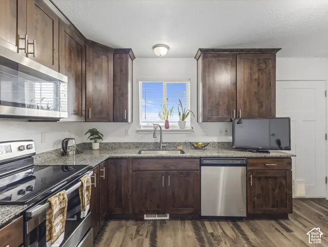 Kitchen featuring wood-finish flooring, granite-style countertops, stainless steel appliances, dark wood cabinetry, and a stainless steel sink with a gooseneck faucet