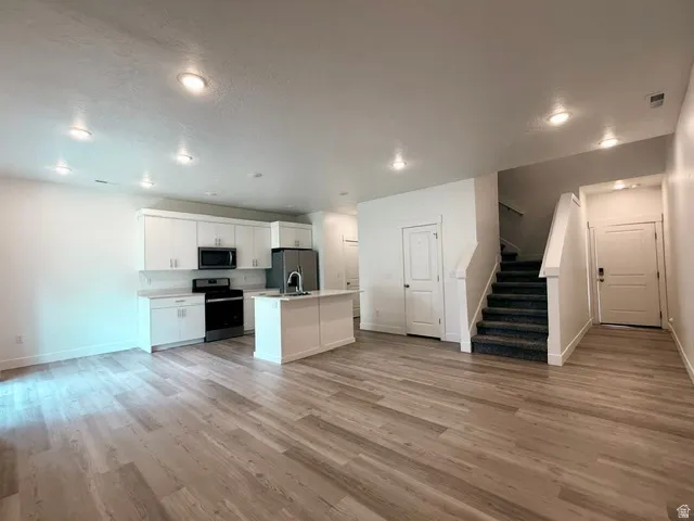 Open-concept living and kitchen area featuring white cabinetry, stainless steel appliances, wood-finish flooring throughout, recessed ceiling lighting, and direct access to a staircase