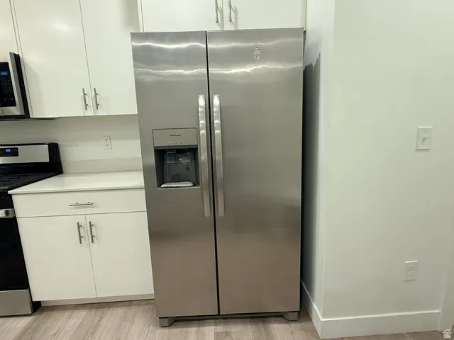 Stainless steel side-by-side refrigerator, white cabinetry, integrated black range, light-toned countertops, and wood-finish flooring