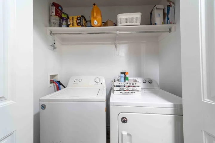 Dedicated laundry room featuring side-by-side washer and dryer appliances, overhead storage shelving with ample capacity for household supplies and linens, and bright, clean white finishes throughout