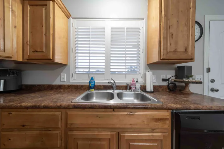 Wood-finish cabinetry, double basin stainless steel sink, black dishwasher, and white window shutters