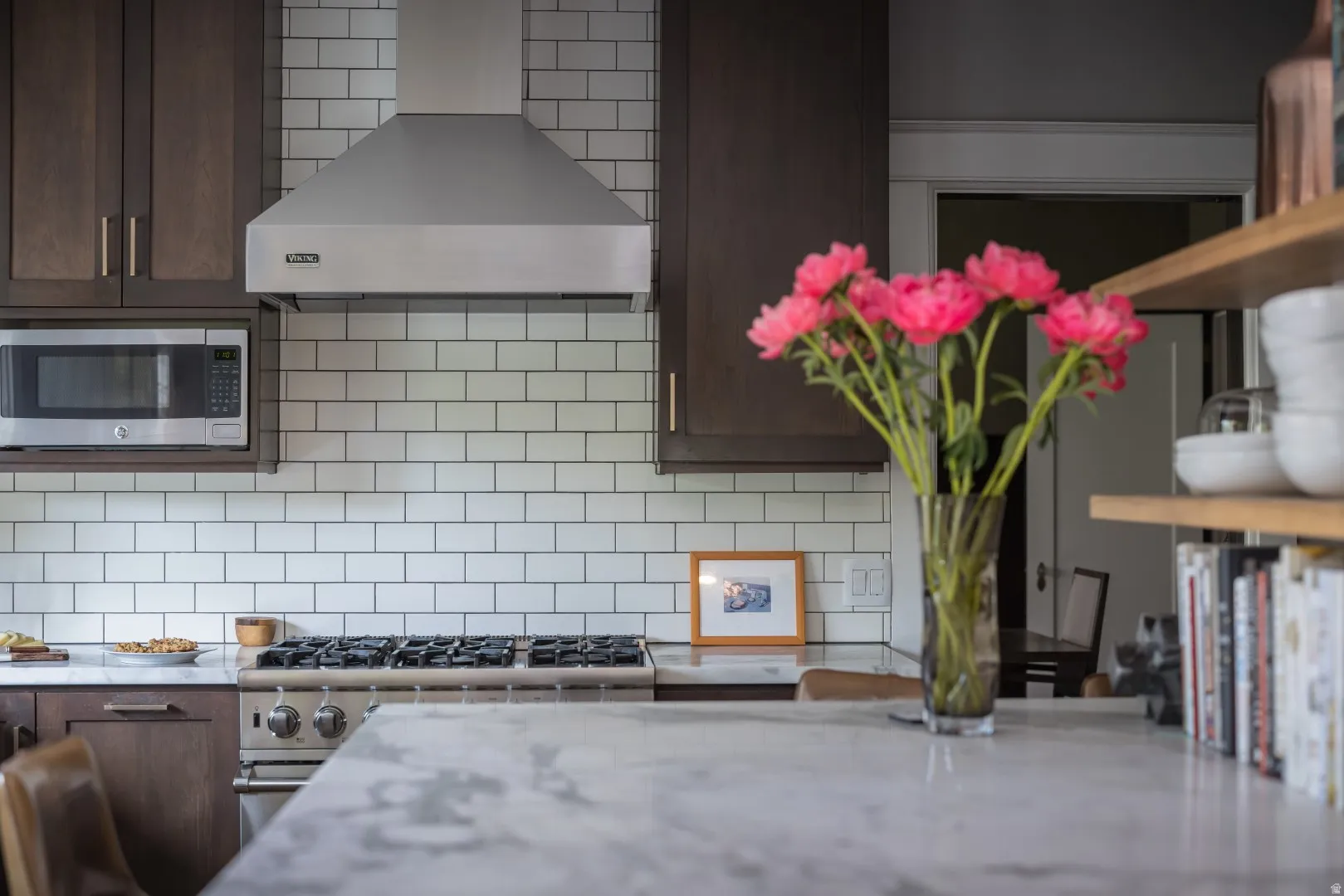 Modern kitchen featuring stainless steel gas cooktop with professional-grade range hood, white subway tile backsplash, dark cabinetry with brushed metal hardware, and a light stone countertop at the waterfall island