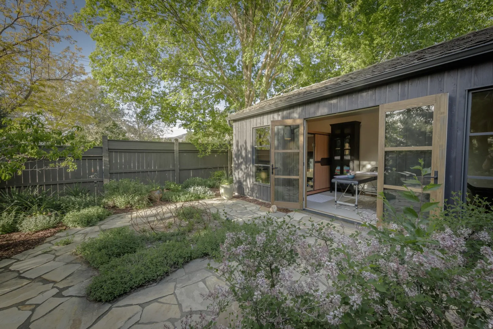 Detached structure with vertical wood siding and double glass-paneled doors, complemented by a flagstone patio and surrounding landscaping