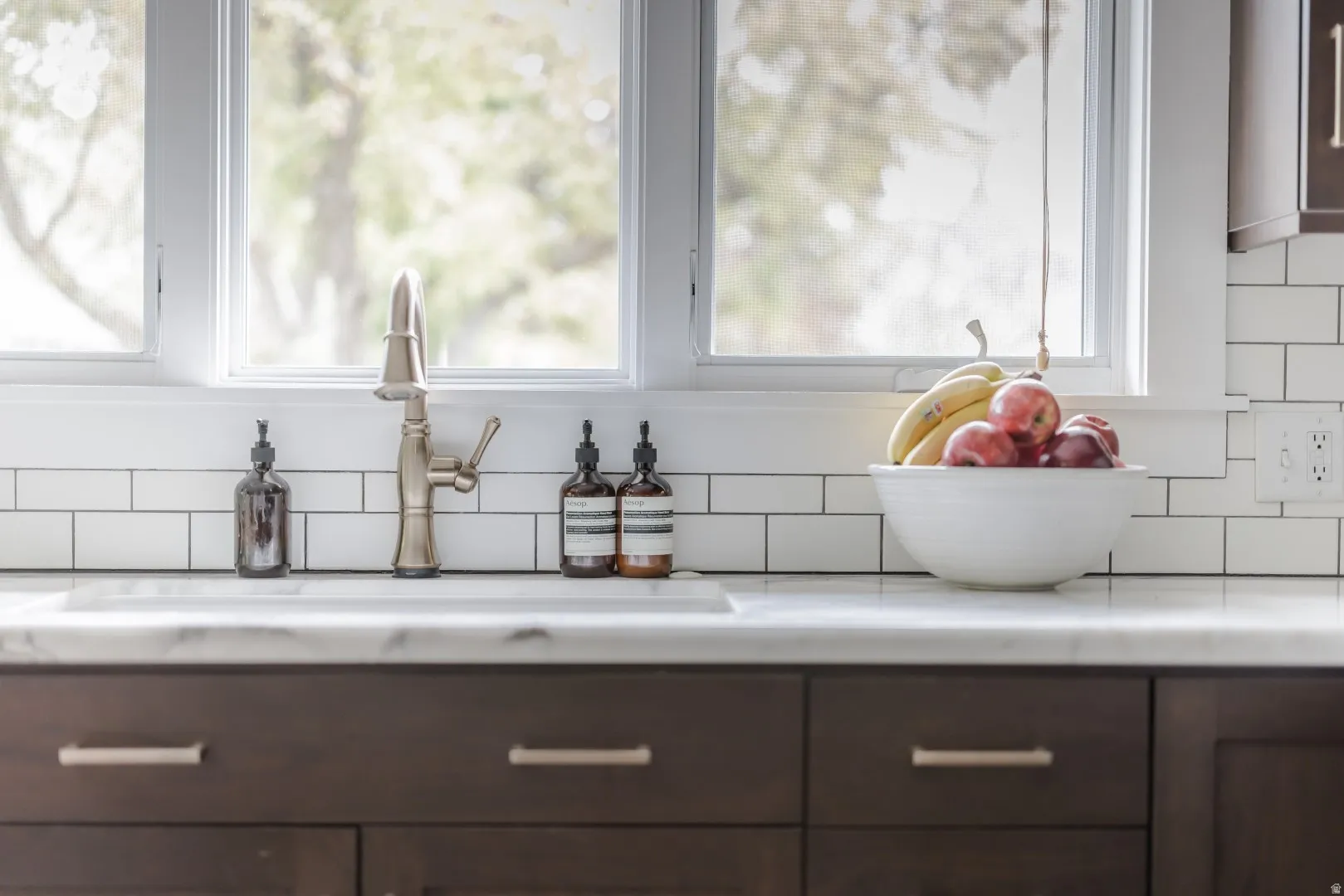 Under-mount sink with a brushed nickel faucet, white subway tile backsplash, white window trim, and dark wood-finish cabinetry