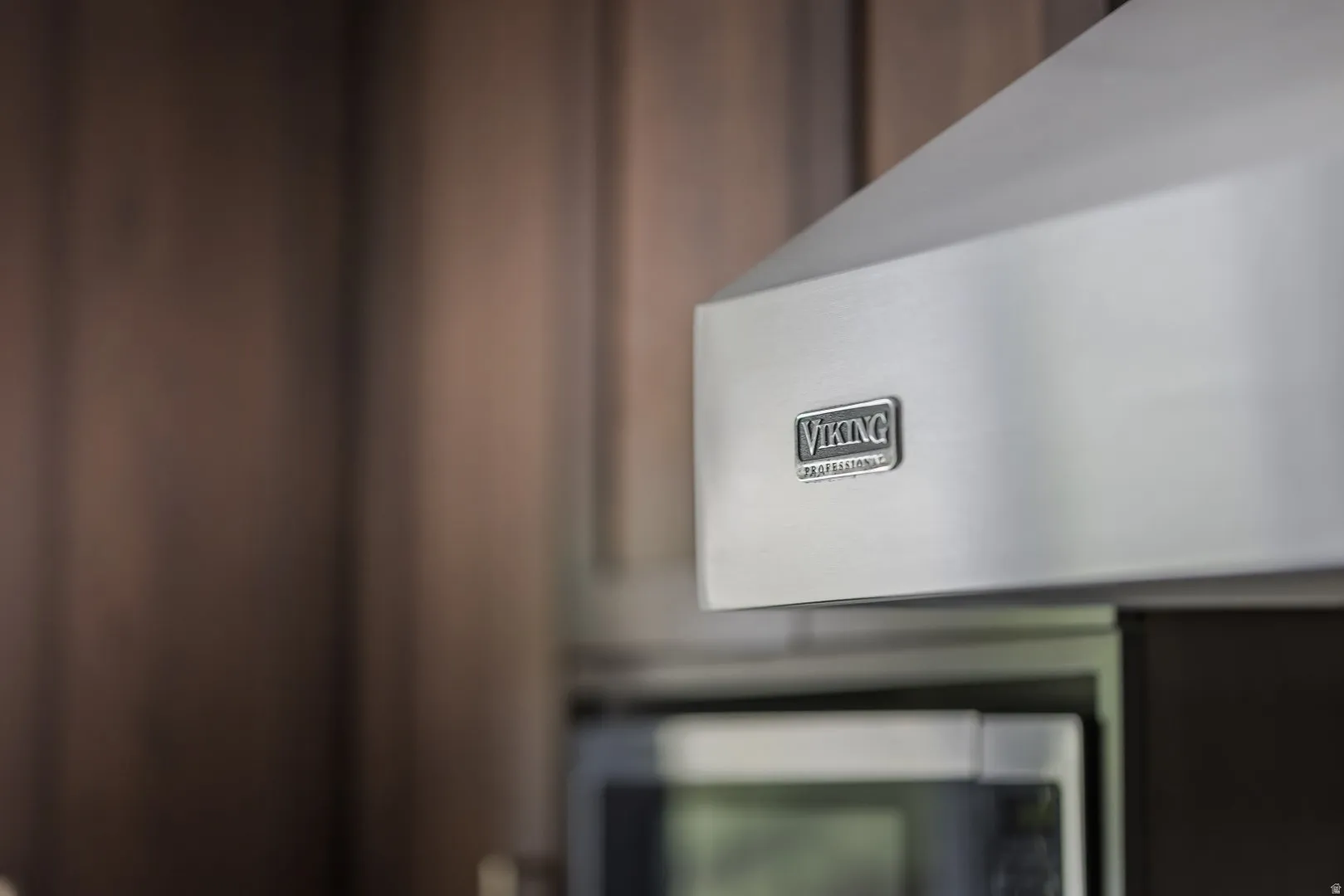 Stainless steel range hood with a prominent brand emblem, integrated above a microwave oven, set against dark wood-finish cabinetry