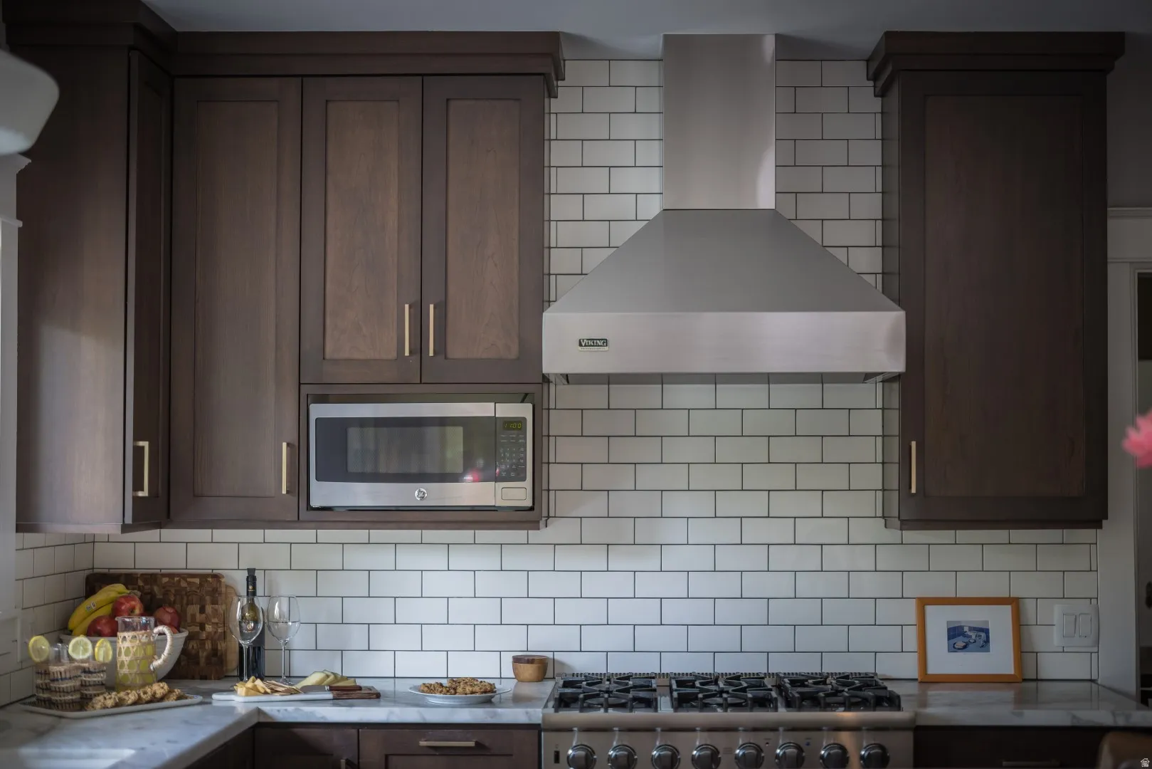 Dark wood-finish cabinetry, stainless steel microwave, stainless steel range hood, white subway tile backsplash, and a gas range with multiple burners