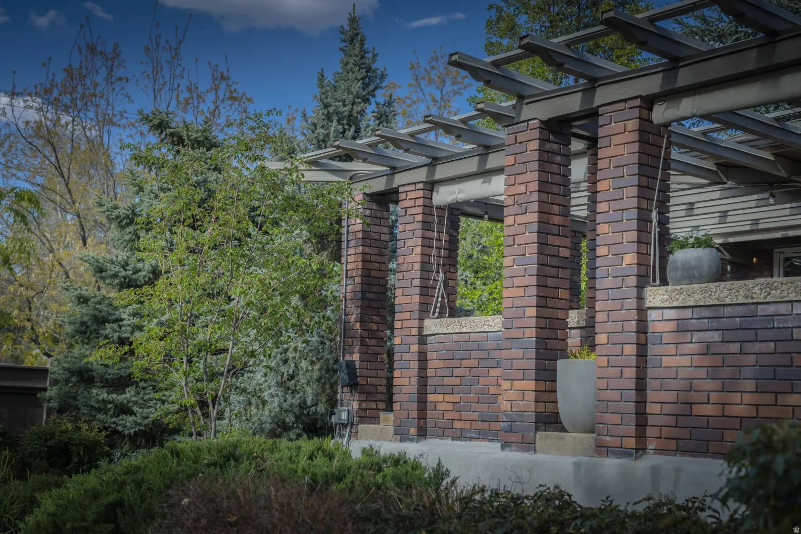Brick columns supporting a wood pergola structure, with surrounding mature trees and shrubs