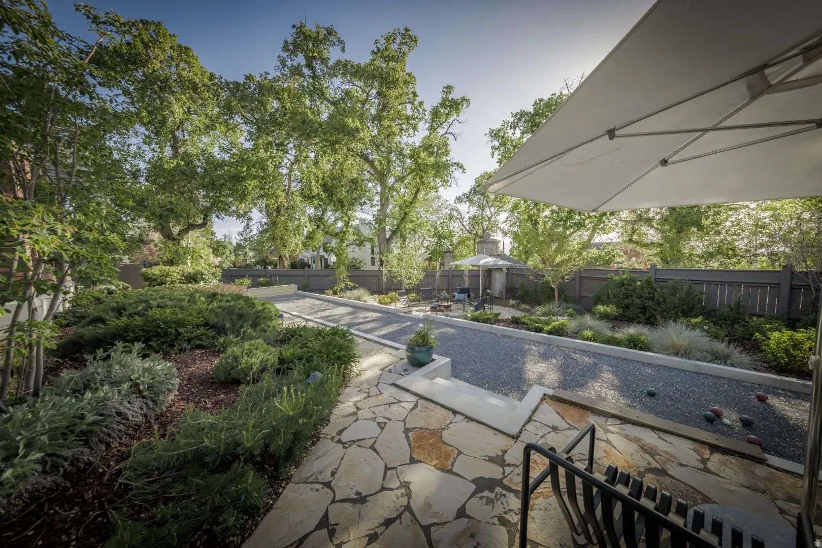 Sophisticated outdoor living space featuring a linear water feature with stone coping, expansive flagstone patio with mixed-stone detailing, mature landscape plantings including shrubs and trees, retractable shade structure, and seamless integration with