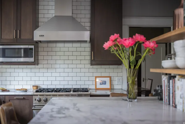 Modern kitchen featuring stainless steel gas cooktop with professional-grade range hood, white subway tile backsplash, dark cabinetry with brushed metal hardware, and a light stone countertop at the waterfall island
