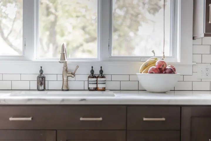 Under-mount sink with a brushed nickel faucet, white subway tile backsplash, white window trim, and dark wood-finish cabinetry