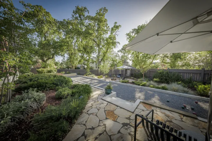 Sophisticated outdoor living space featuring a linear water feature with stone coping, expansive flagstone patio with mixed-stone detailing, mature landscape plantings including shrubs and trees, retractable shade structure, and seamless integration with
