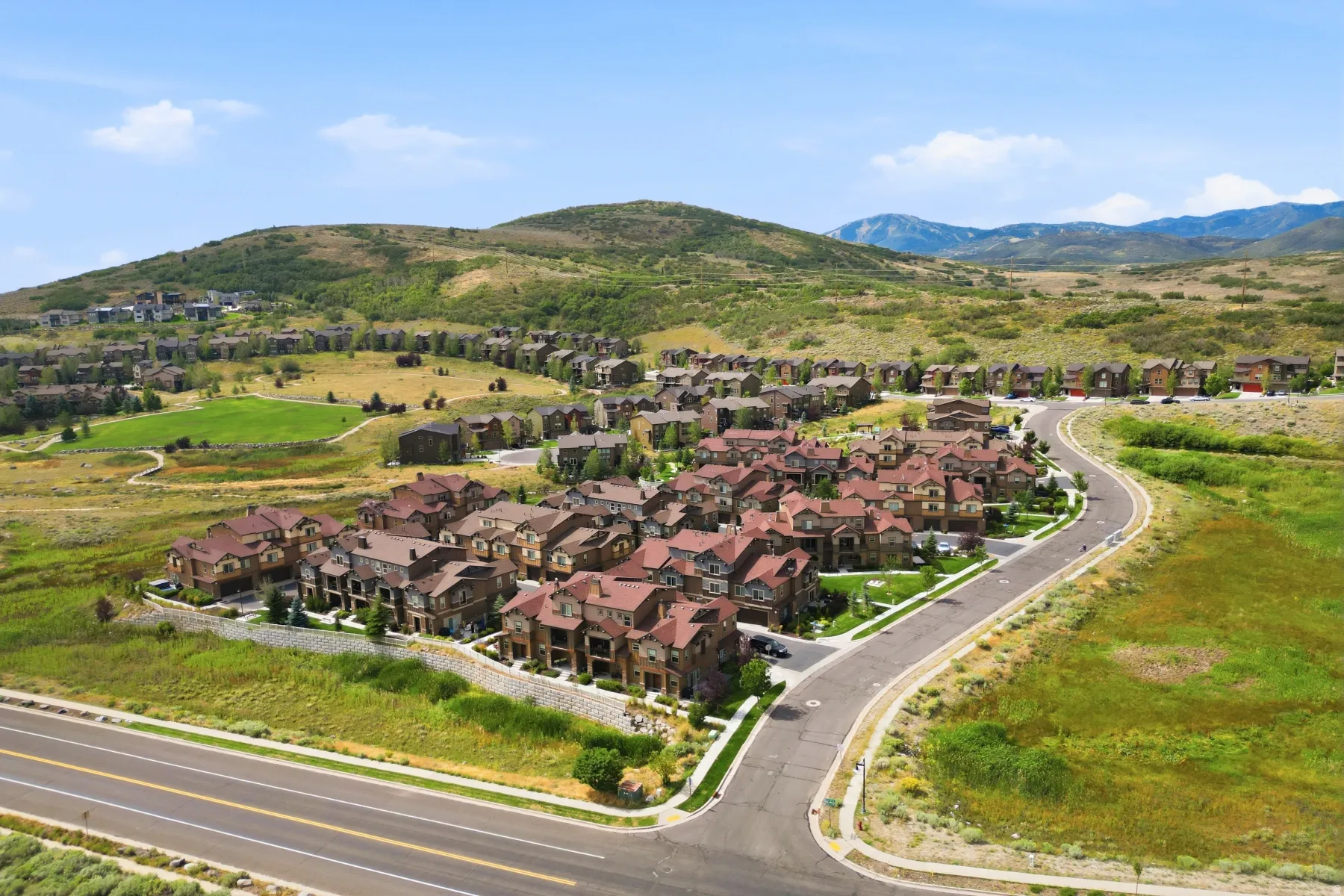 Residential community with multi-level properties and red-tile roofs, bordered by paved roads and natural landscaping, set against a backdrop of rolling hills and distant mountains