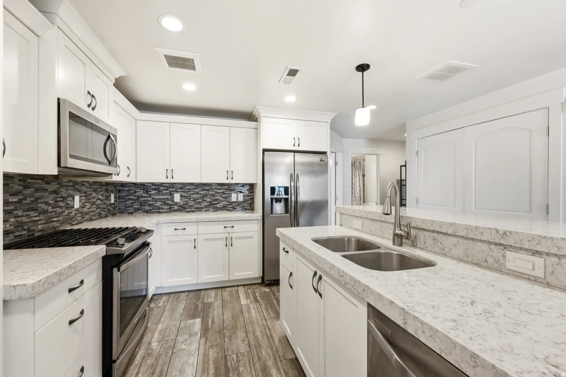 White cabinetry with black hardware, stainless steel appliances, dual basin sink, wood-finish flooring, and a mosaic tile backsplash