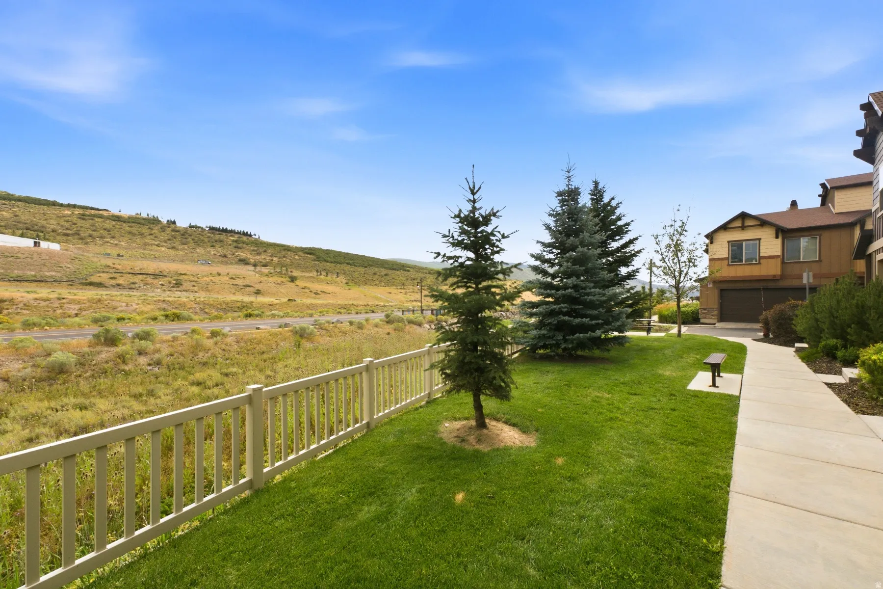 Manicured lawn with mature evergreen trees, a concrete pathway, and a light-colored fence