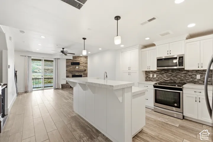 Open-concept layout with wood-finish flooring, white shaker cabinetry, stainless steel appliances, and a stone-clad fireplace