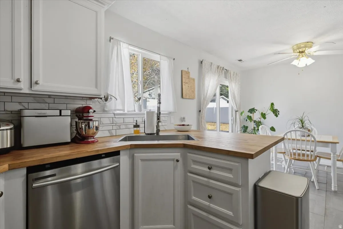 White cabinetry with dark hardware, stainless steel dishwasher, light wood-finish countertops, subway tile backsplash, and a corner sink