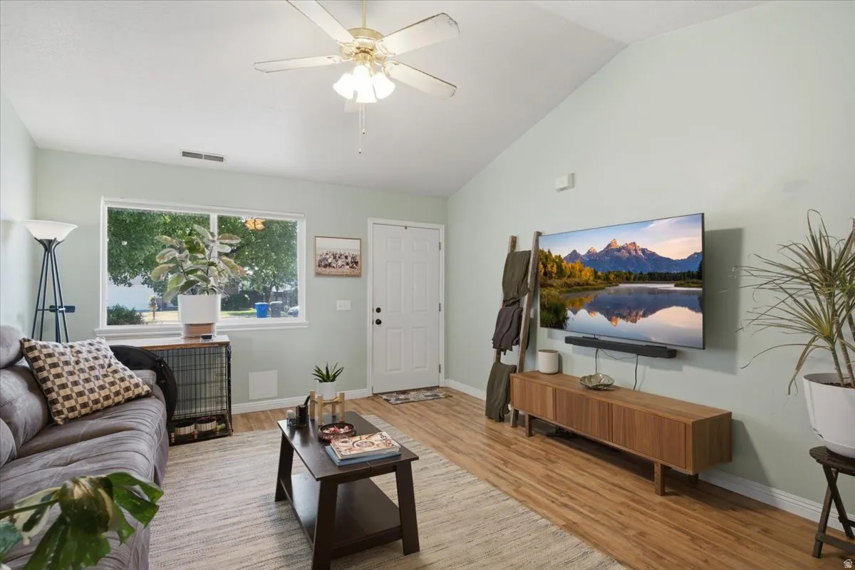 Bright, open living room showcasing warm wood-finish flooring, vaulted ceilings with ceiling fan and integrated lighting, and large picture window with natural light