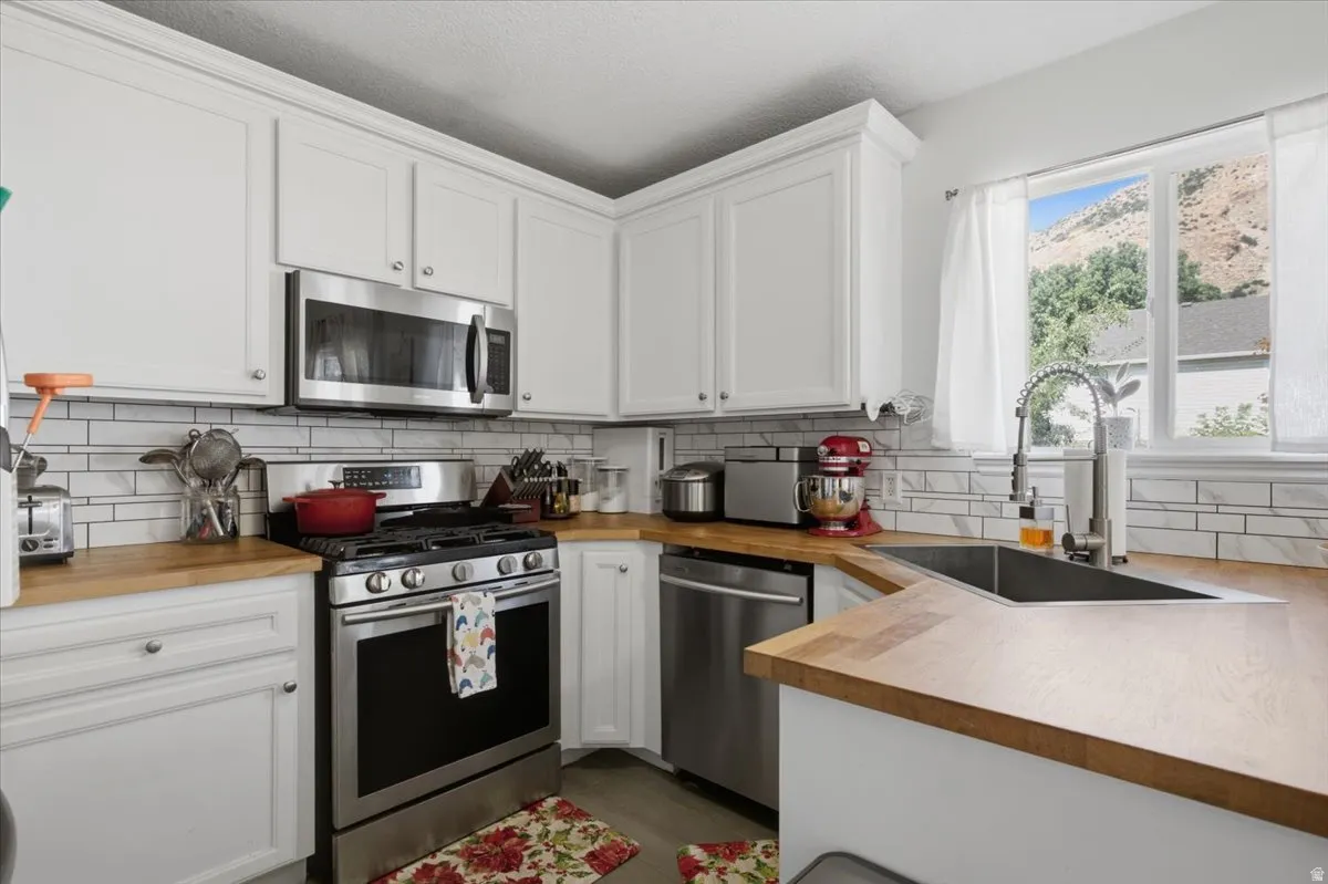 White cabinetry with crown molding, stainless steel appliances, wood-finish countertops, subway tile backsplash, and a corner sink