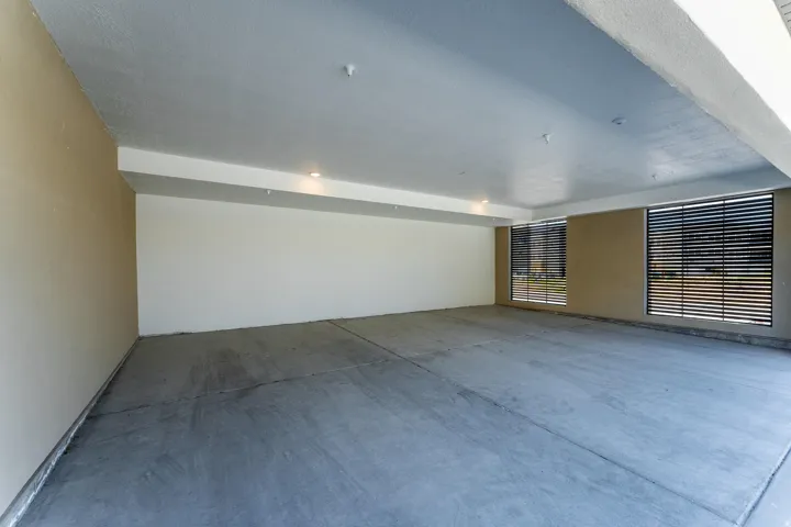 Spacious covered parking area featuring a concrete floor, two large windows with horizontal slat covers, recessed lighting, and neutral-toned walls