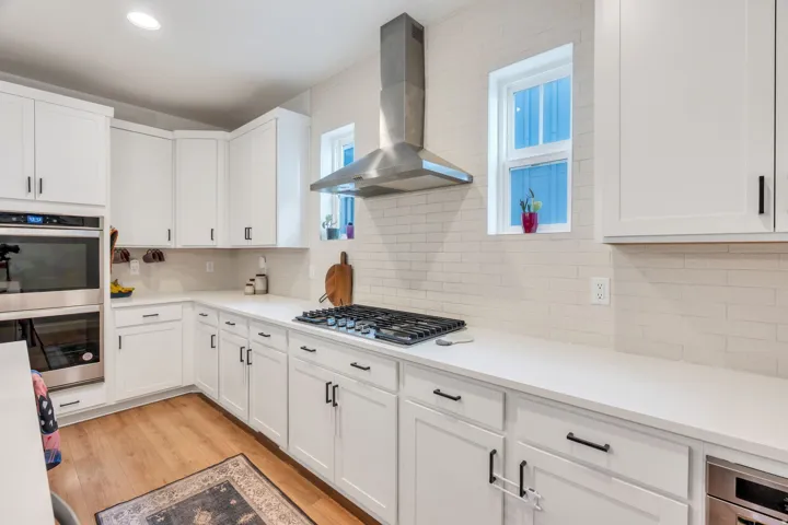 White cabinetry with dark hardware, built-in stainless steel oven and microwave, gas cooktop with stainless steel range hood, light-toned backsplash, and wood-finish flooring