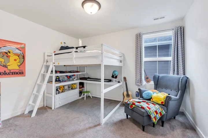 Versatile bedroom featuring a white metal loft bed with integrated shelving and workspace below, carpeted flooring, large double-hung window with coordinating curtains, and a comfortable reading chair positioned near natural light