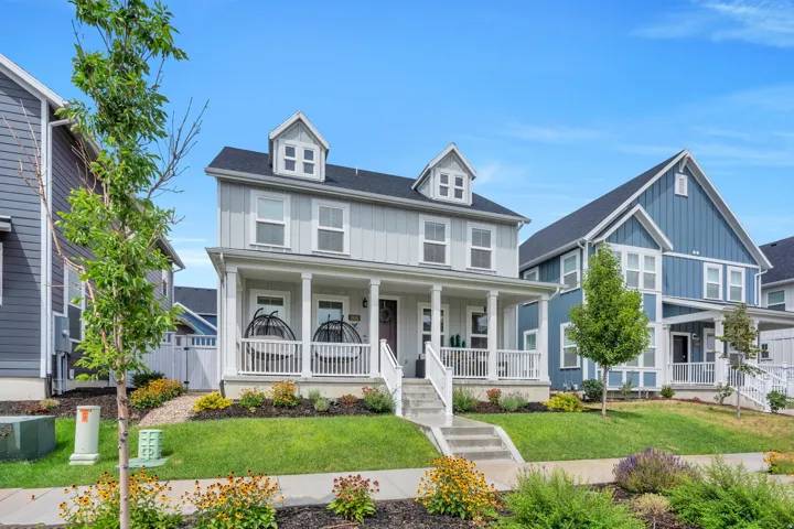 Two-story residence featuring a covered front porch, dormer windows, a dark shingle roof, and a mature landscaped front yard