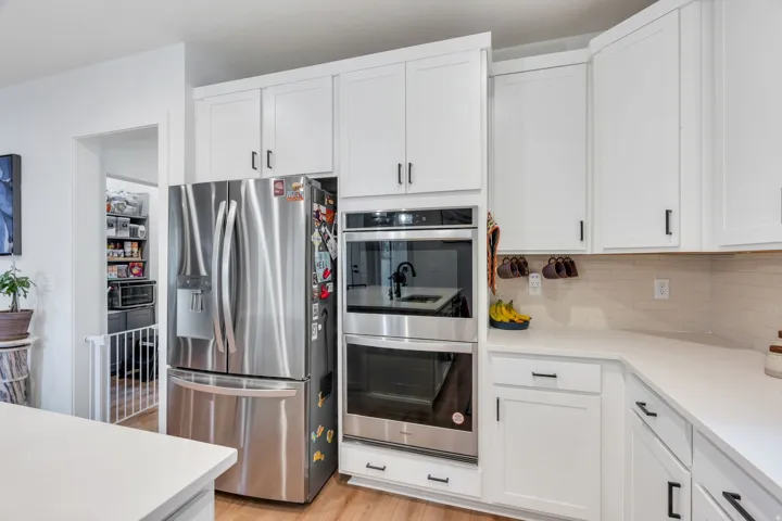Stainless steel refrigerator, built-in double ovens, white shaker cabinetry, light-toned backsplash, and wood-finish flooring